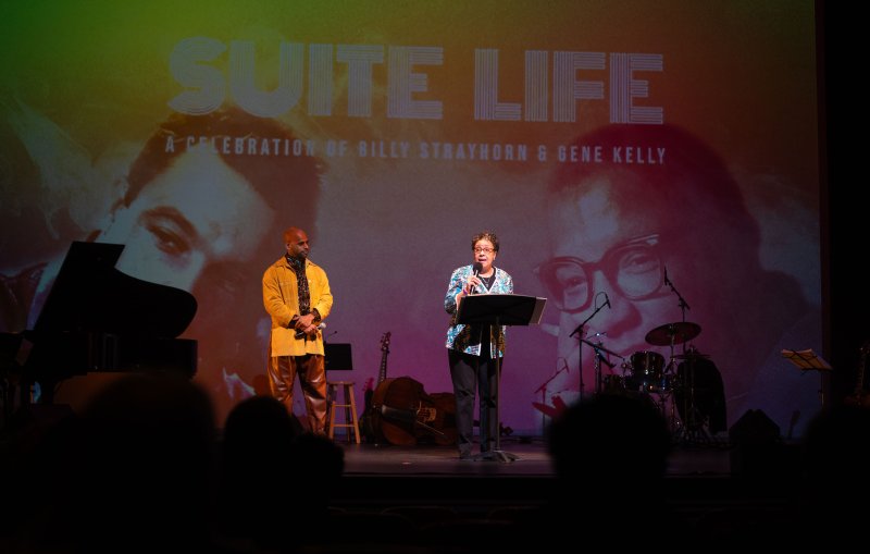 A woman speaks at a podium on a stage with a man, piano, and instruments, in front of a screen displaying "SUITE LIFE.