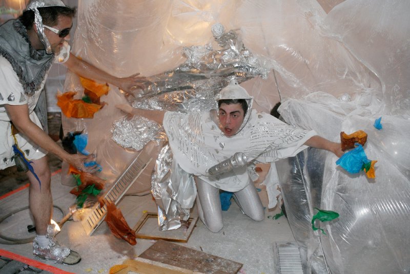 Performance art with two people in shiny, futuristic costumes amidst clear plastic and crumpled silver foil.
