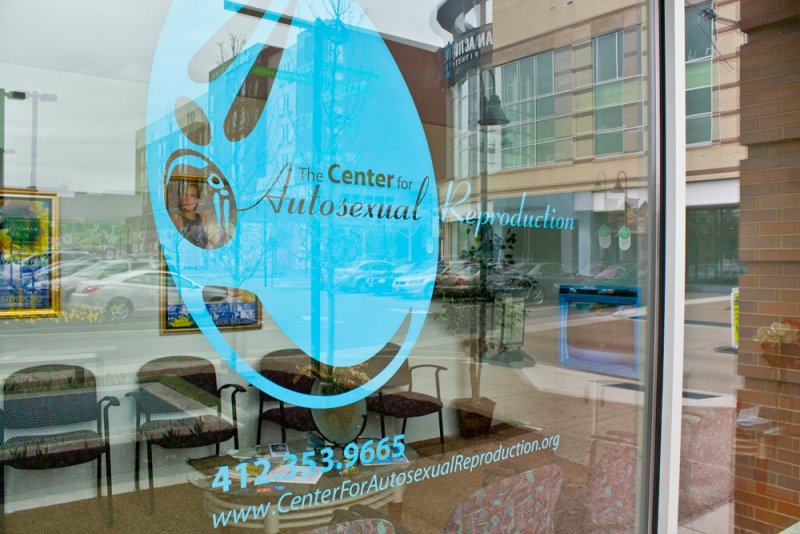Shop window view with blue logo for "The Center for Autosexual Reproduction" overlaid on waiting room interior and street reflections. Chairs, framed art, and contact information visible.