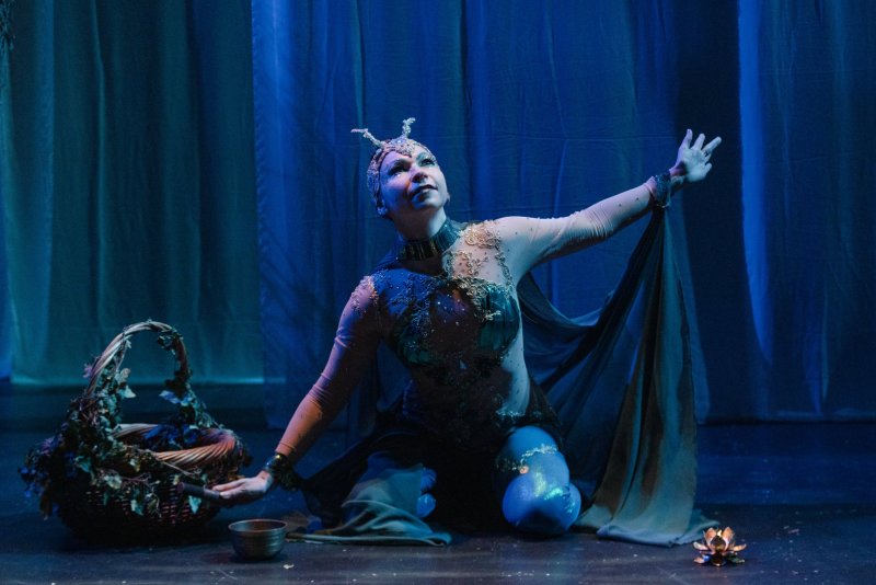 A dancer in an insect-like costume, kneeling on a stage with arms outstretched, under blue and purple lighting.
