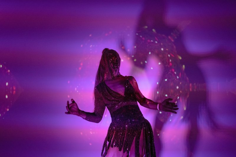 Dancer in a sequined dress, seen from behind, illuminated by purple and pink light with scattered bright dots, creating a blurred shadow.