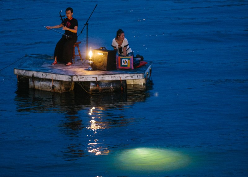 Two people on a wooden raft at dusk, one playing an erhu, the other sitting. A lantern glows, reflecting on dark blue water.