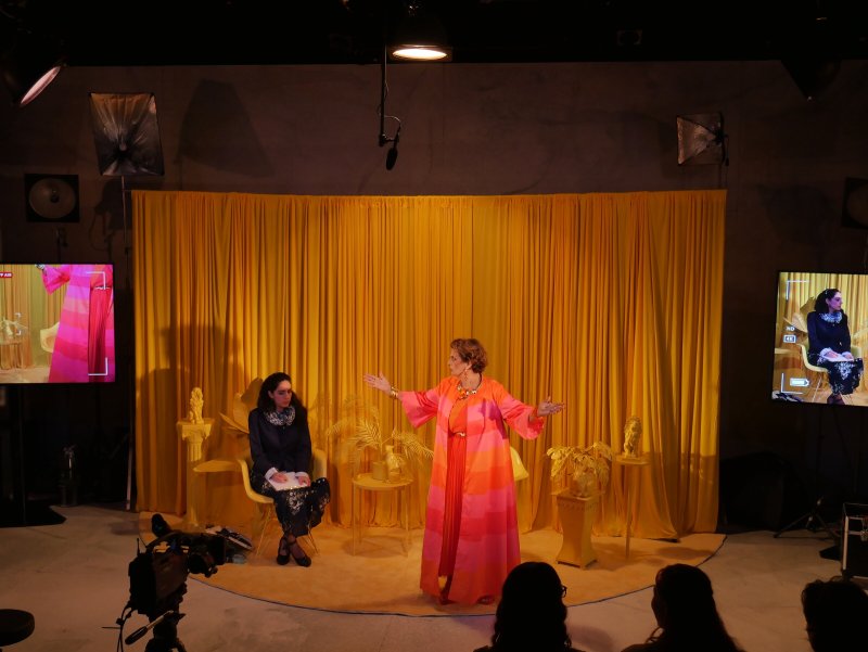 Two women on a stage with a bright yellow curtain, one in a pink and orange dress gesticulating, the other seated.