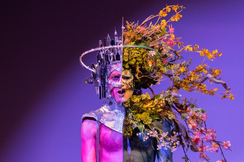 Performer Darrell Thorne in a split costume: left is silver with a cityscape crown, right is organic with branches and flowers.
