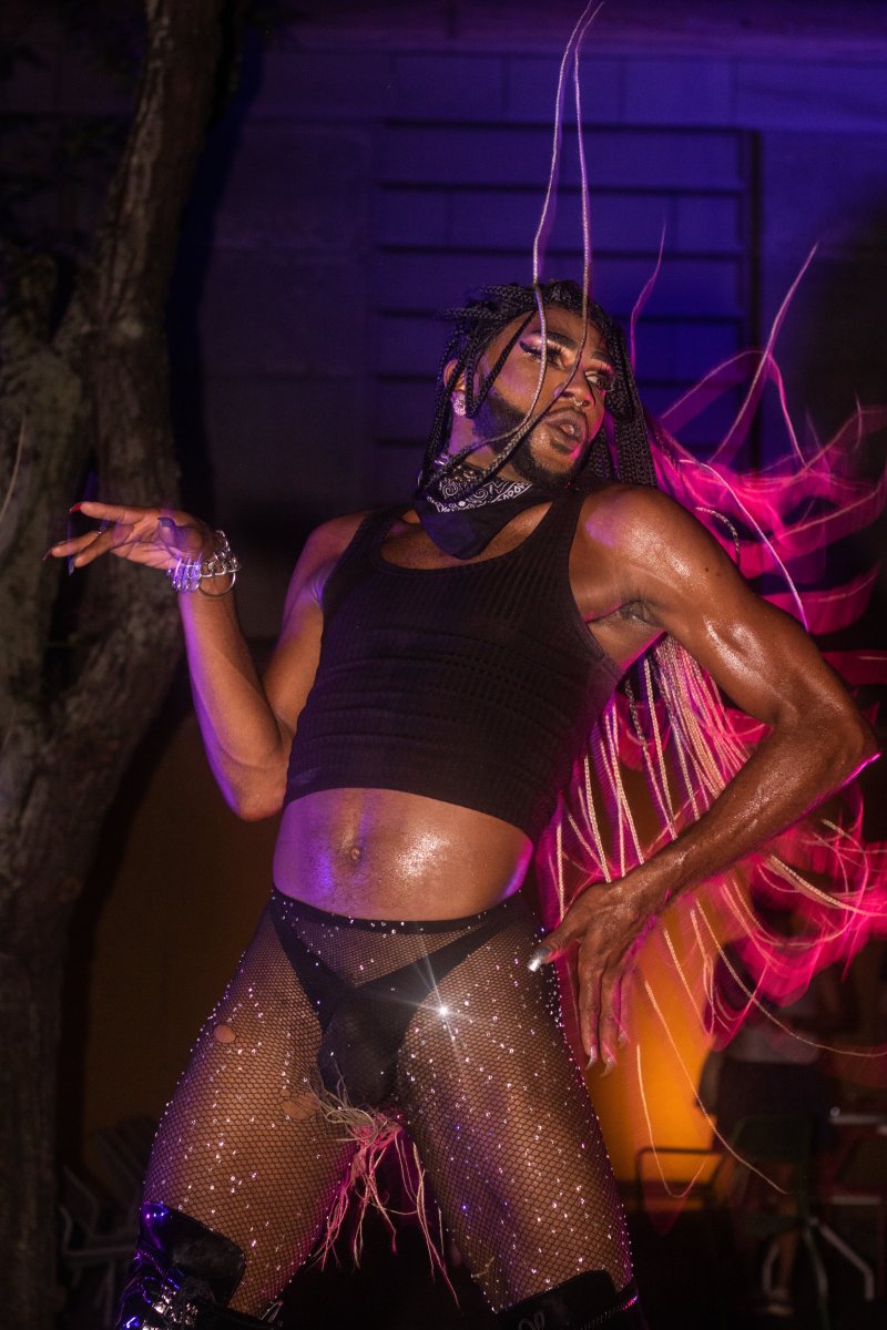 Performer in black tank top and fishnet tights with rhinestones poses under purple and pink lights. Braided hair with silver strands, bandana necklace, and platform boots complete the look.