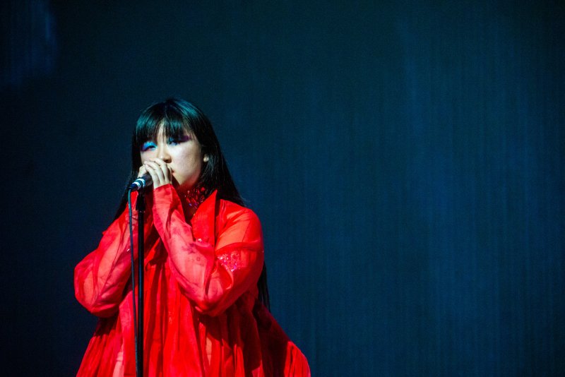 Woman with long dark hair, wearing a flowing red dress, singing into a microphone against a dark blue background.