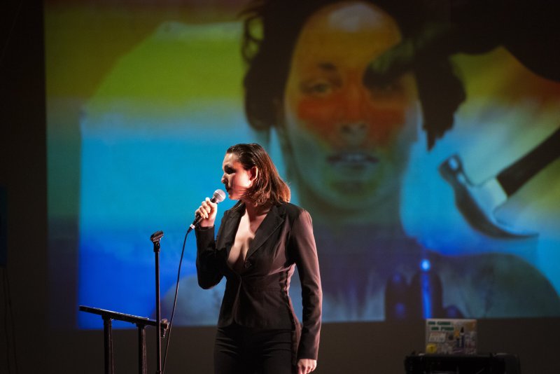 Performance art with a singer in a black suit against a projected, blurred face in blue, green, and orange light.