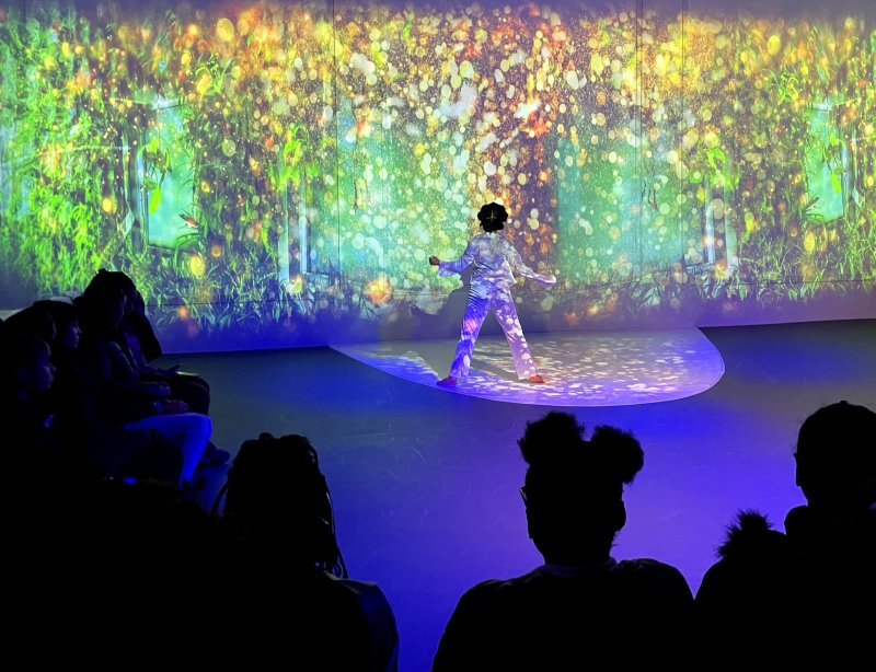 Performance art installation with a figure in a patterned suit center stage, bathed in blue light. A lush, green and gold abstract projection serves as the backdrop, viewed by a silhouetted audience.