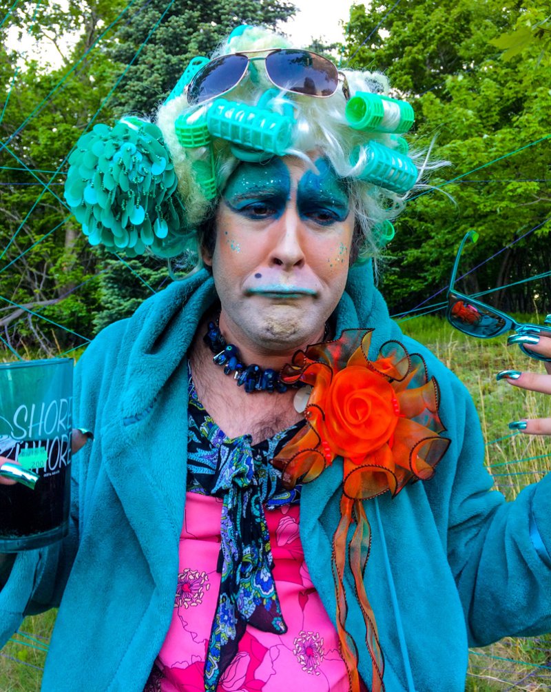 Person in teal robe, pink dress, and blue makeup with hair curlers and sunglasses, holding a drink.