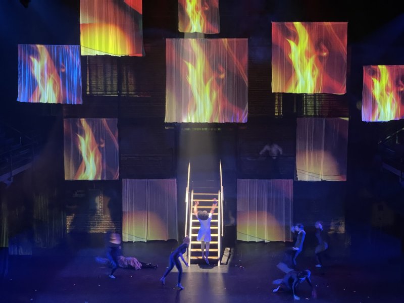 Stage performance with dancers in blue light. Projected flame patterns on hanging translucent squares. A woman climbs a ladder center stage.