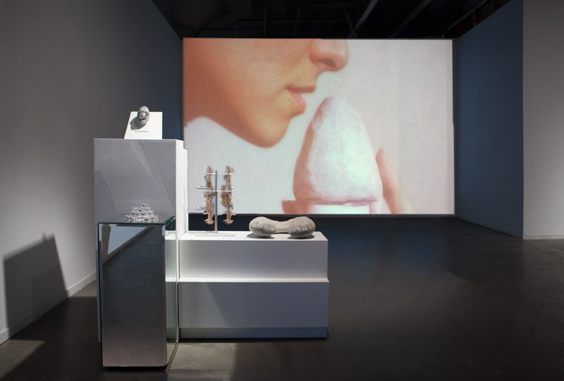 Gallery installation with a large projection of a person smelling a white object, and pedestals displaying small, abstract sculptures.
