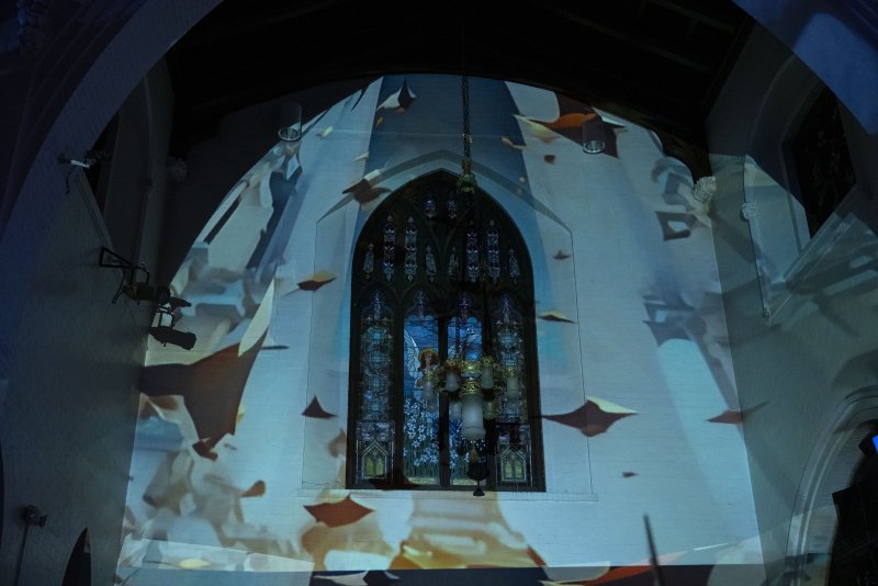 Projected blue light and abstract shapes on a white church wall with a stained-glass window in the center.