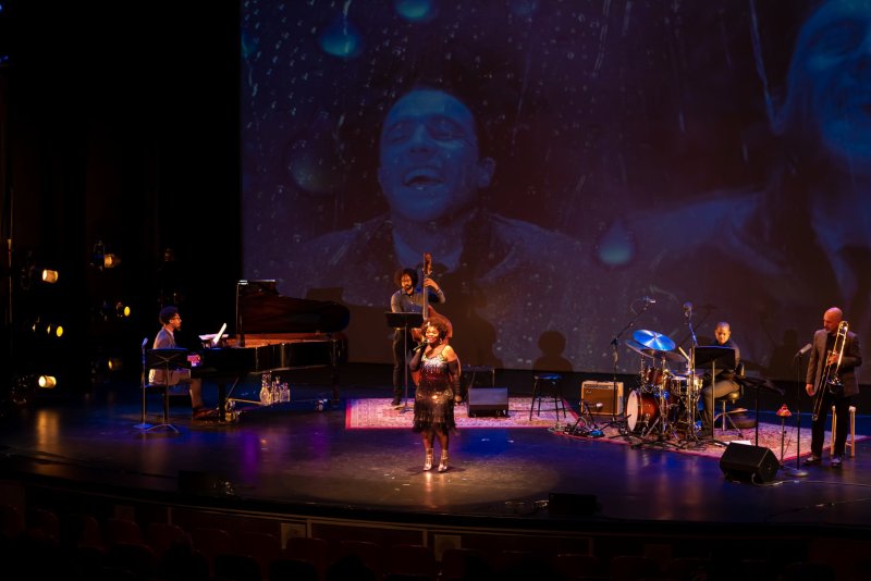 A singer in a sparkly dress performs on stage with a pianist, bassist, drummer, and trombonist, against a projected blue image of a man's face.