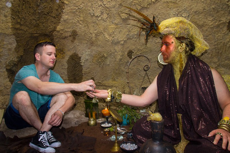 Two figures in a cave-like setting. One man in casual clothes offers an object to a figure adorned in gold body paint, a feathered headdress, and draped in a brown, sequined fabric. Candles and ritualistic objects are arranged on the ground.
