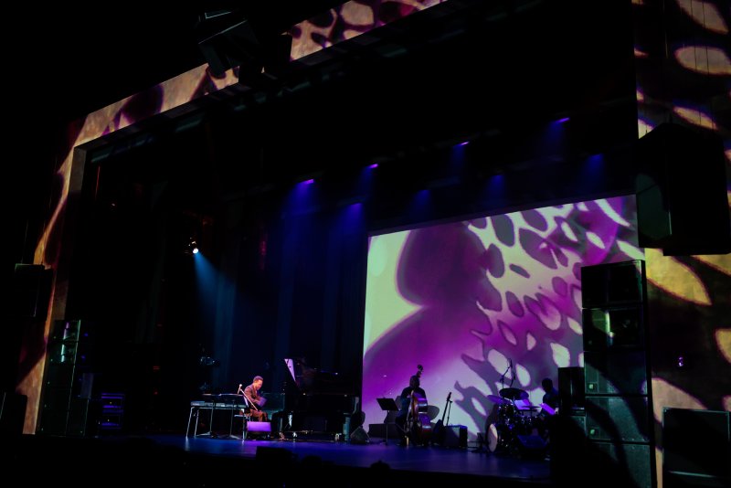 Dark stage with musicians, a pianist, and a bassist, under purple and blue lights, with abstract purple and green projections.