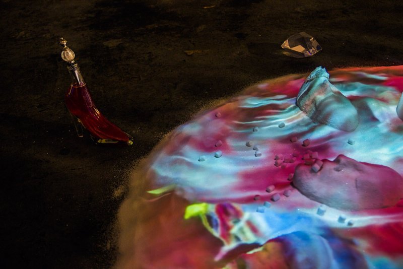 Installation view featuring a red glass shoe-shaped bottle, a diamond, and rock-like forms on a surface with swirling pink and blue light patterns, scattered with small cubes.