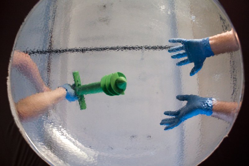 Two gloved hands inside a clear, textured sphere, reaching for a green, propeller-like object.