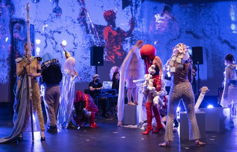 Performance art with costumed figures on stage, blue projected digital art on background wall.