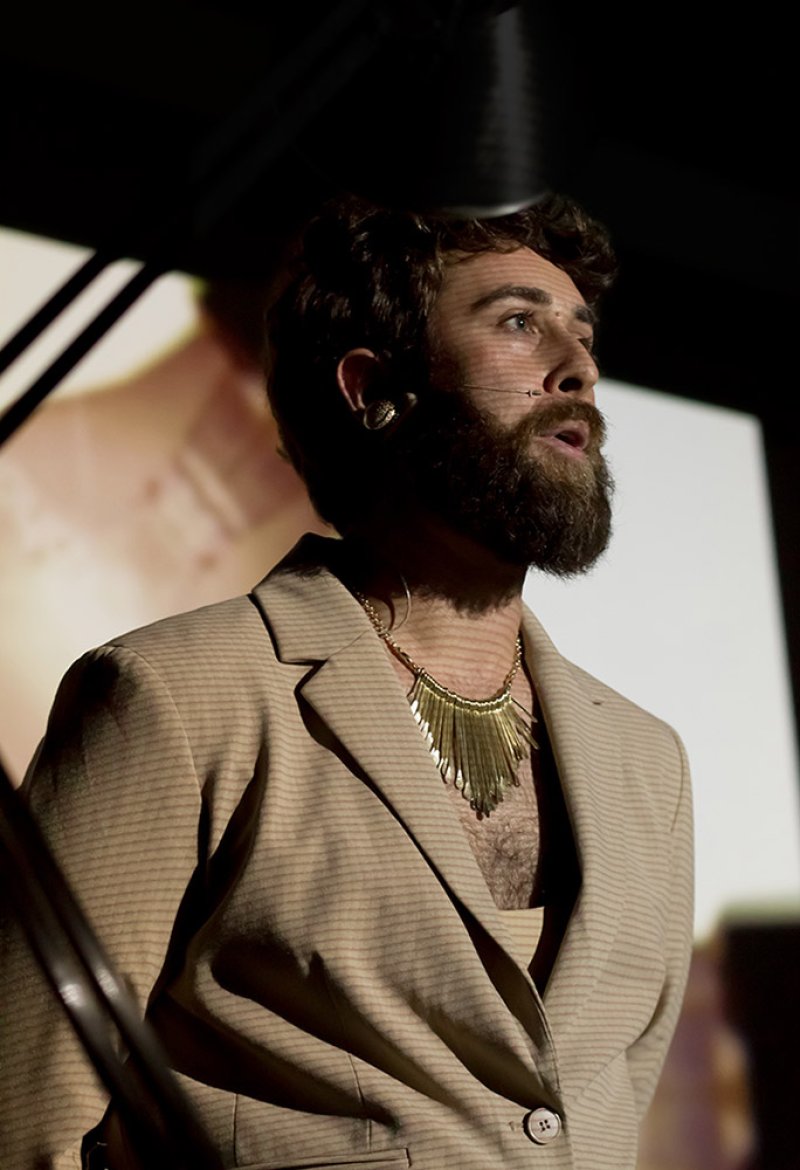 Man with beard, gold necklace, and beige blazer looking right, with a projected image in the background.