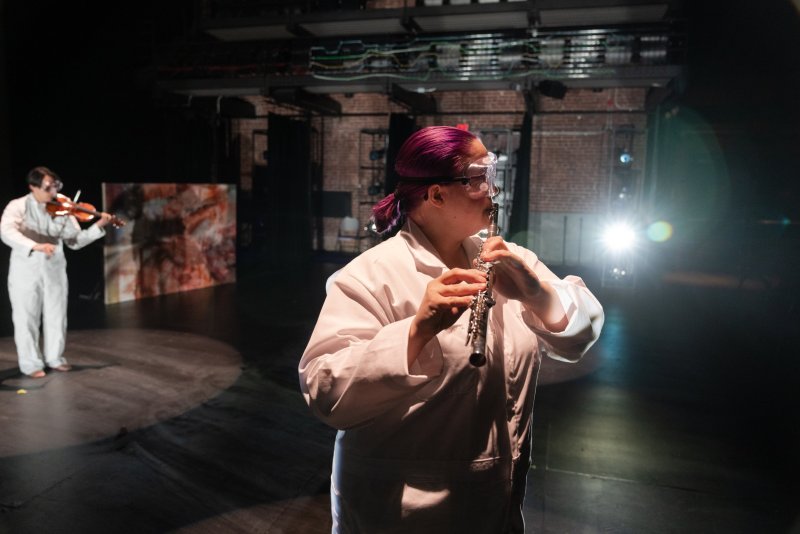 Woman in white lab coat and goggles plays flute on dark stage, another person in white plays violin in background.