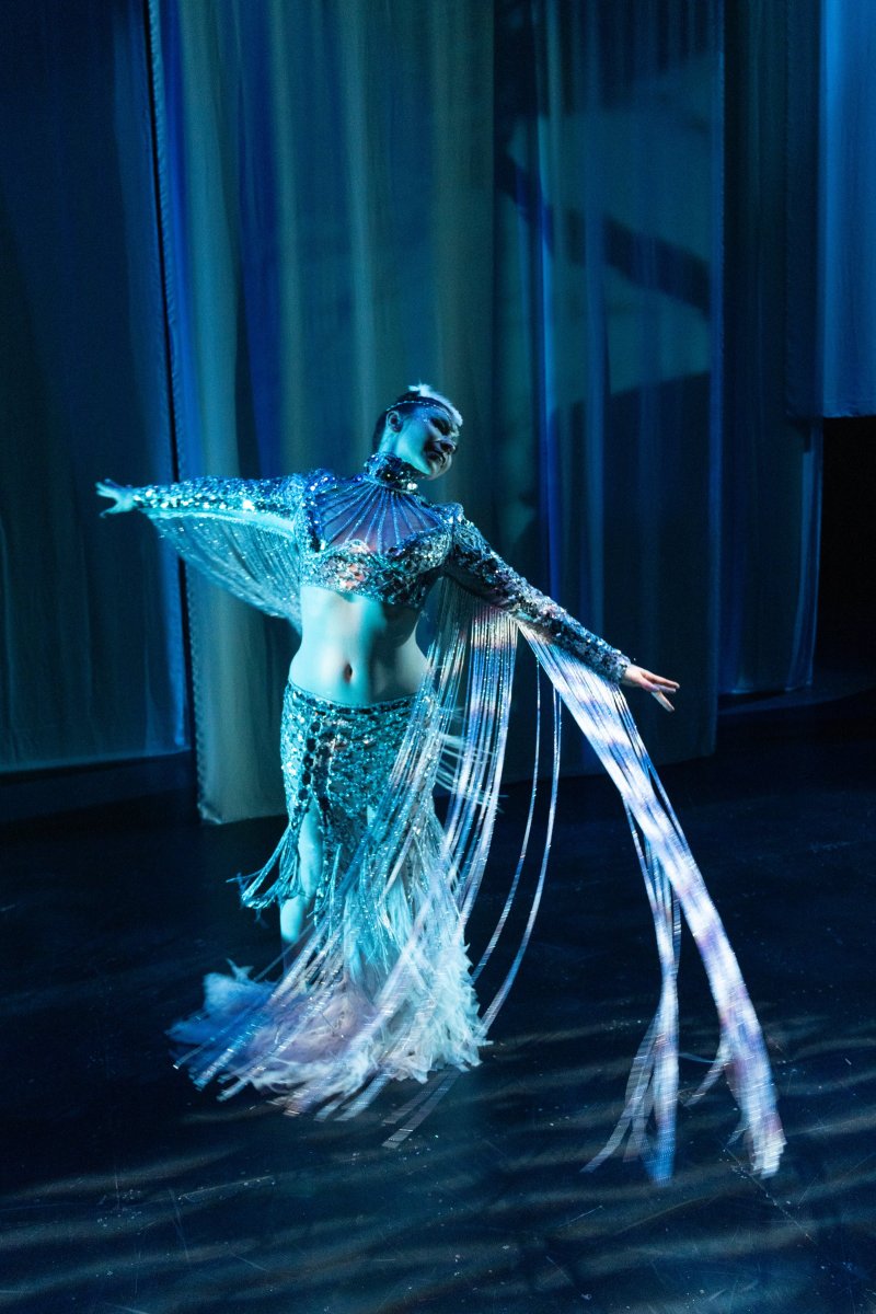 A dancer in a shimmering silver costume with long, flowing sleeves and a skirt, illuminated by blue light on a dark stage.