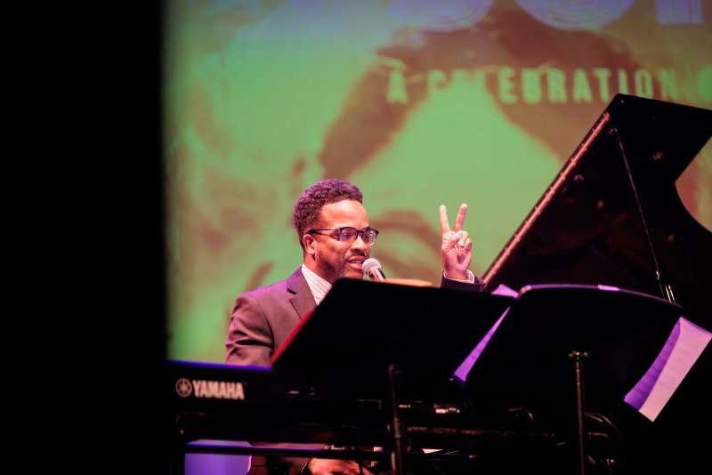 Man in suit and glasses at a Yamaha grand piano, holding up two fingers, with a green and orange blurred background.
