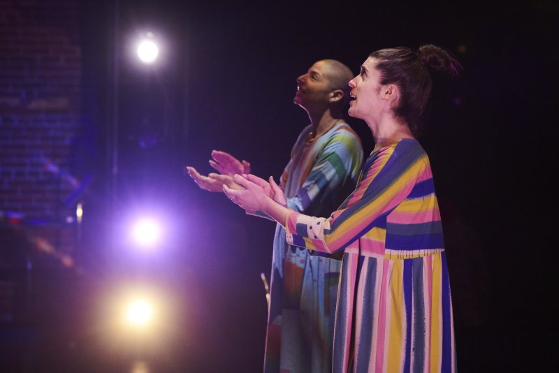 Two figures, lit by spotlights, face upwards with hands raised. They wear brightly striped, colorful clothing in vertical patterns. Background is dark and indistinct.