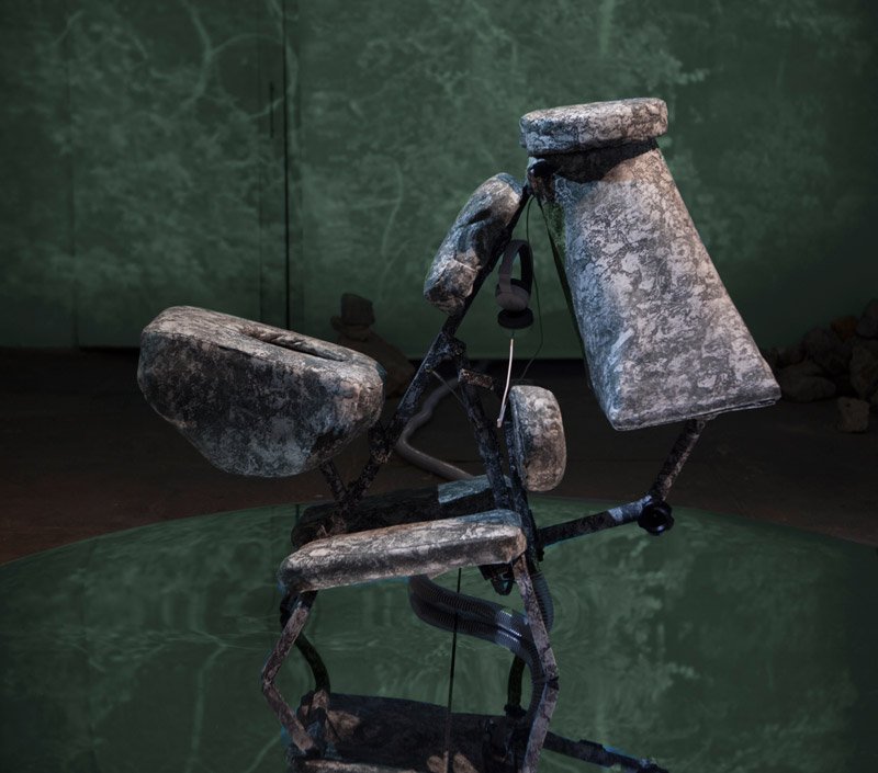Sculptural chair made of gray, textured stone-like cushions on a dark metal frame, with headphones hanging from it, against a green background.