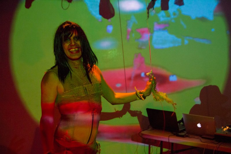Woman with dark hair, wearing a sheer, textured top, holds a puppet against a green and red projected background.