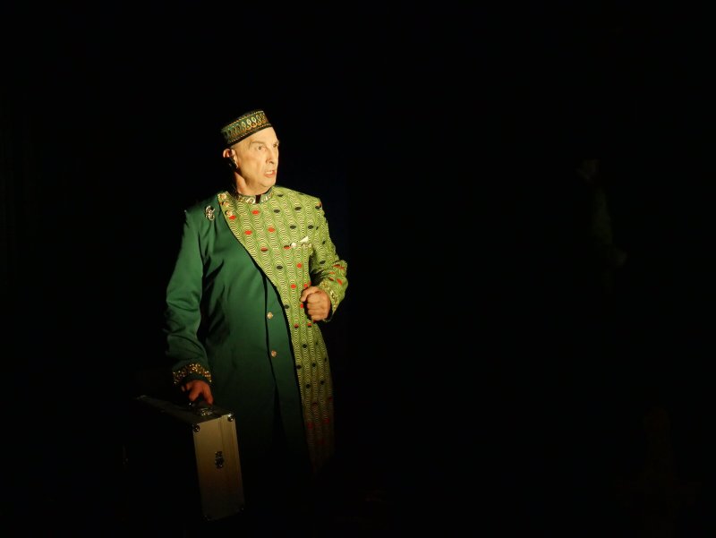 Man in green suit with patterned sash and hat, holding a briefcase, lit from the left against a dark background.