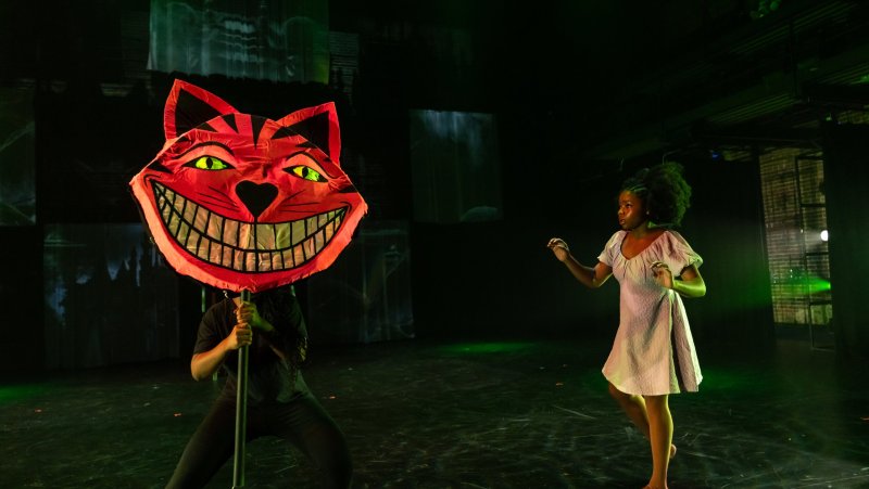 Stage view of a person holding a large red Cheshire Cat head, facing a woman in a white dress, with dark green lighting.