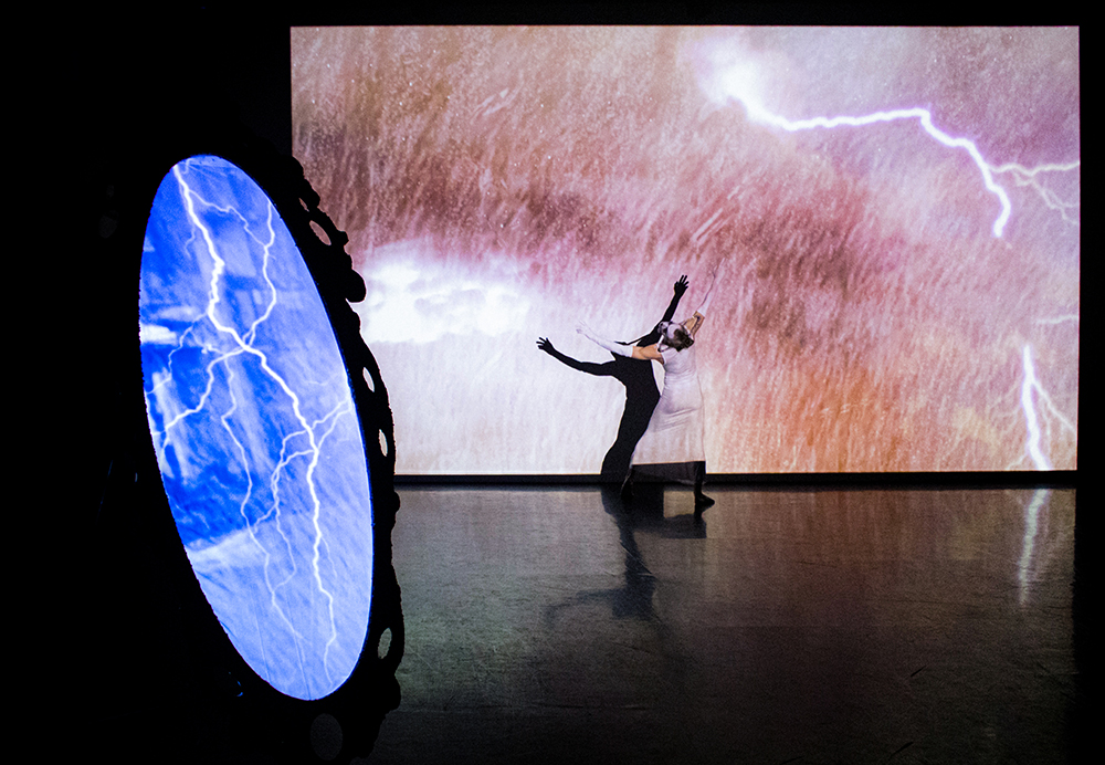 Performance with a dancer in white and black, arms outstretched, before a screen displaying pink lightning.