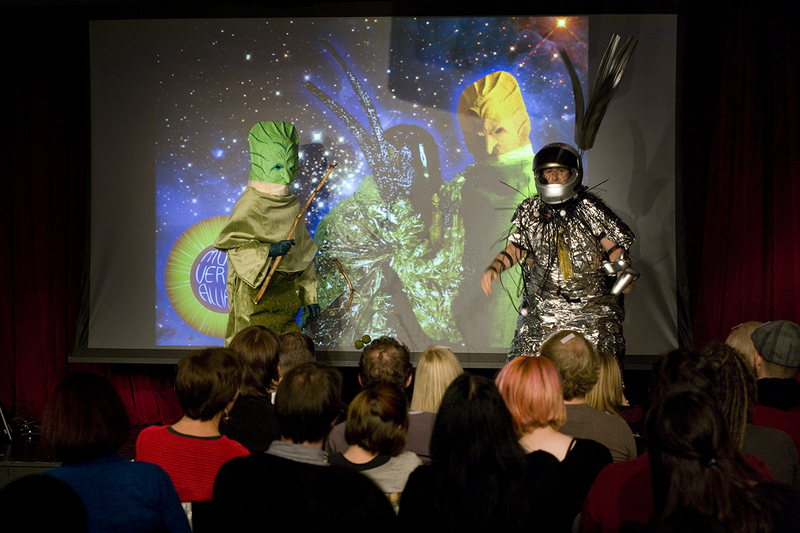 Two performers in alien costumes on stage, one green, one silver, in front of a cosmic projection.